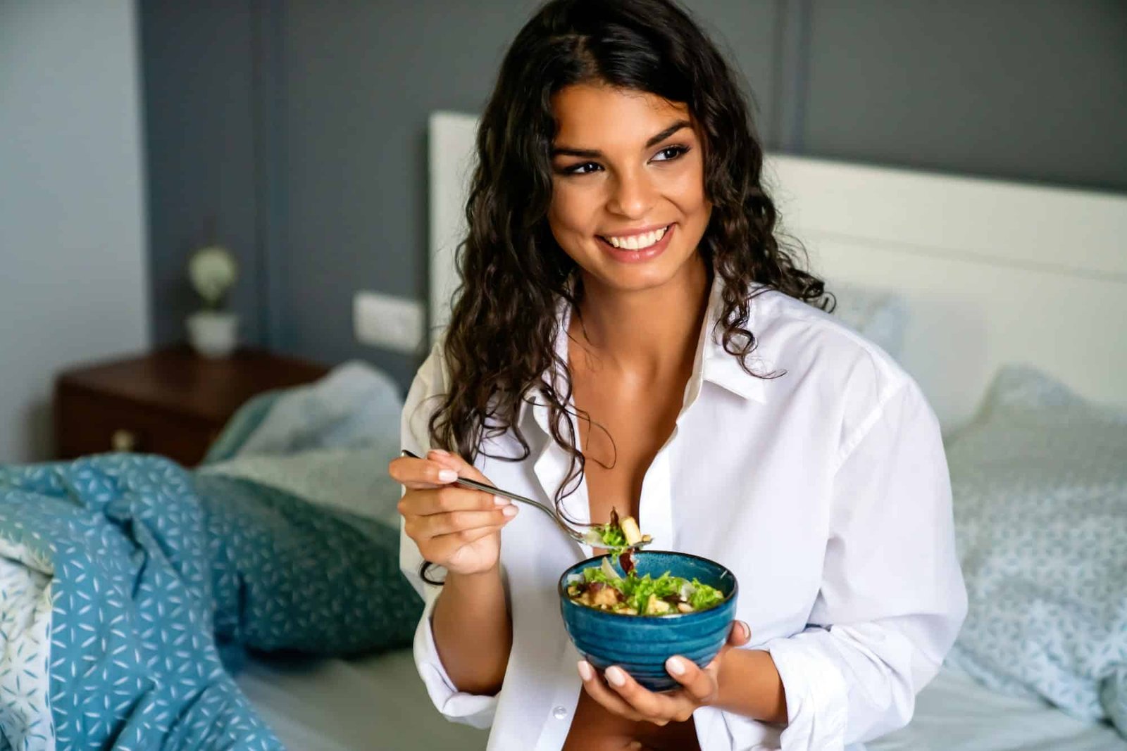 Portrait of a sexy young happy woman eating healthy salad in bedroom.